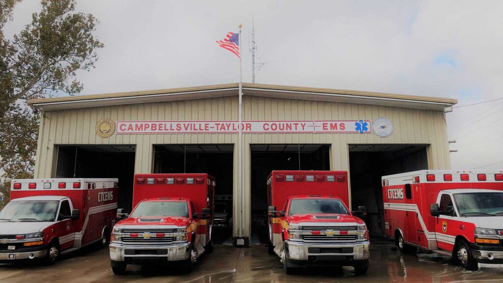 Four Campbellsville–Taylor County EMS ambulances parked in front of the emergency services building with an American flag above.